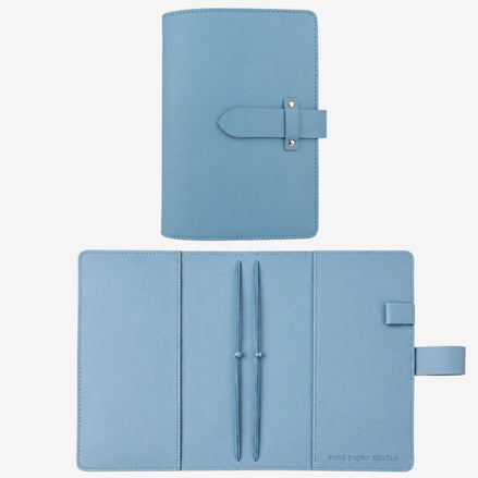 A light blue leather planner cover shown closed at the top and open at the bottom on a white background. The open view reveals two pen loops with pens, a pen slot, and a small brand mark reading 'post paper studio' in the lower right corner.