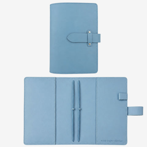A light blue leather planner cover shown closed at the top and open at the bottom on a white background. The open view reveals two pen loops with pens, a pen slot, and a small brand mark reading 'post paper studio' in the lower right corner.