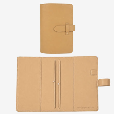 A tan leather planner cover shown closed and open on a light gray background. The closed view shows a strap-and-button closure, and the open view reveals interior pockets, card slots, and a small stitched brand mark reading 'rust paper studio' near the lower right.